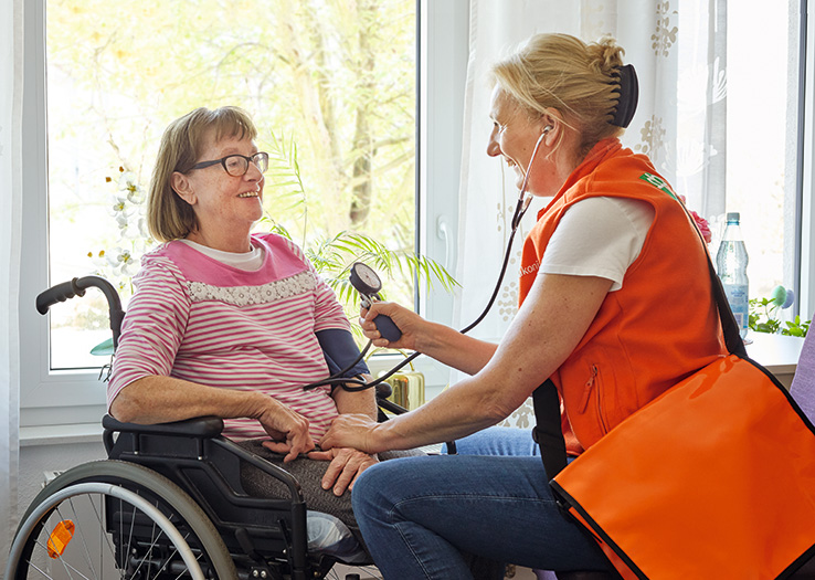 Pflegefachkraft des Ambulanten Dienstes misst Blutdruck bei einer Dame im Rollstuhl
