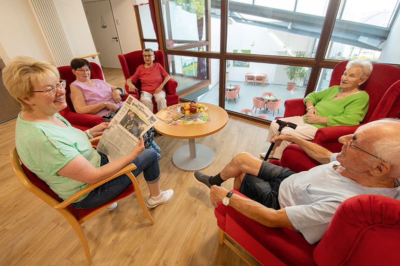 Pflegefachfrau liest Bewohnern aus der Zeitung vor, Blick in die Eingangshalle