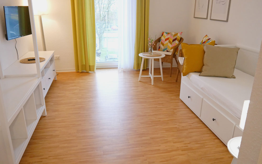 Blick in einen Raum mit Holzfußboden, TV an der linken und Bett an der rechten Wand