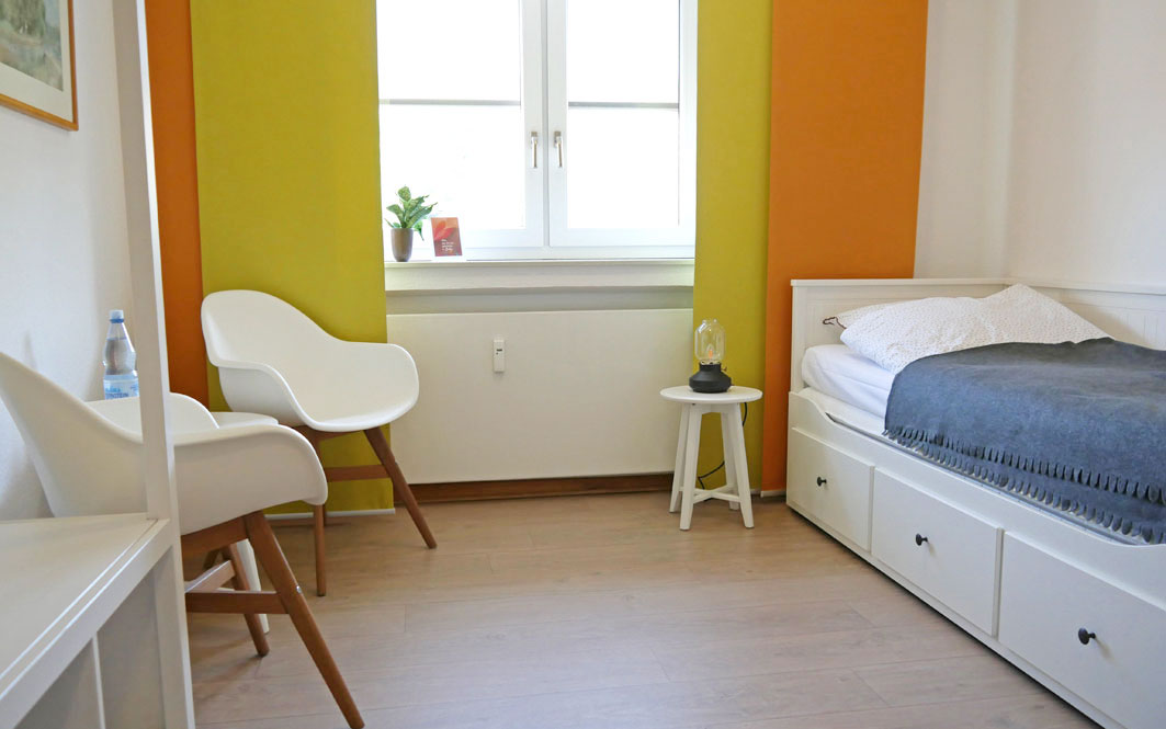 Ein Zimmer mit einem Bett an der rechten Wand und einer kleinen Sitzecke an der linken Wand