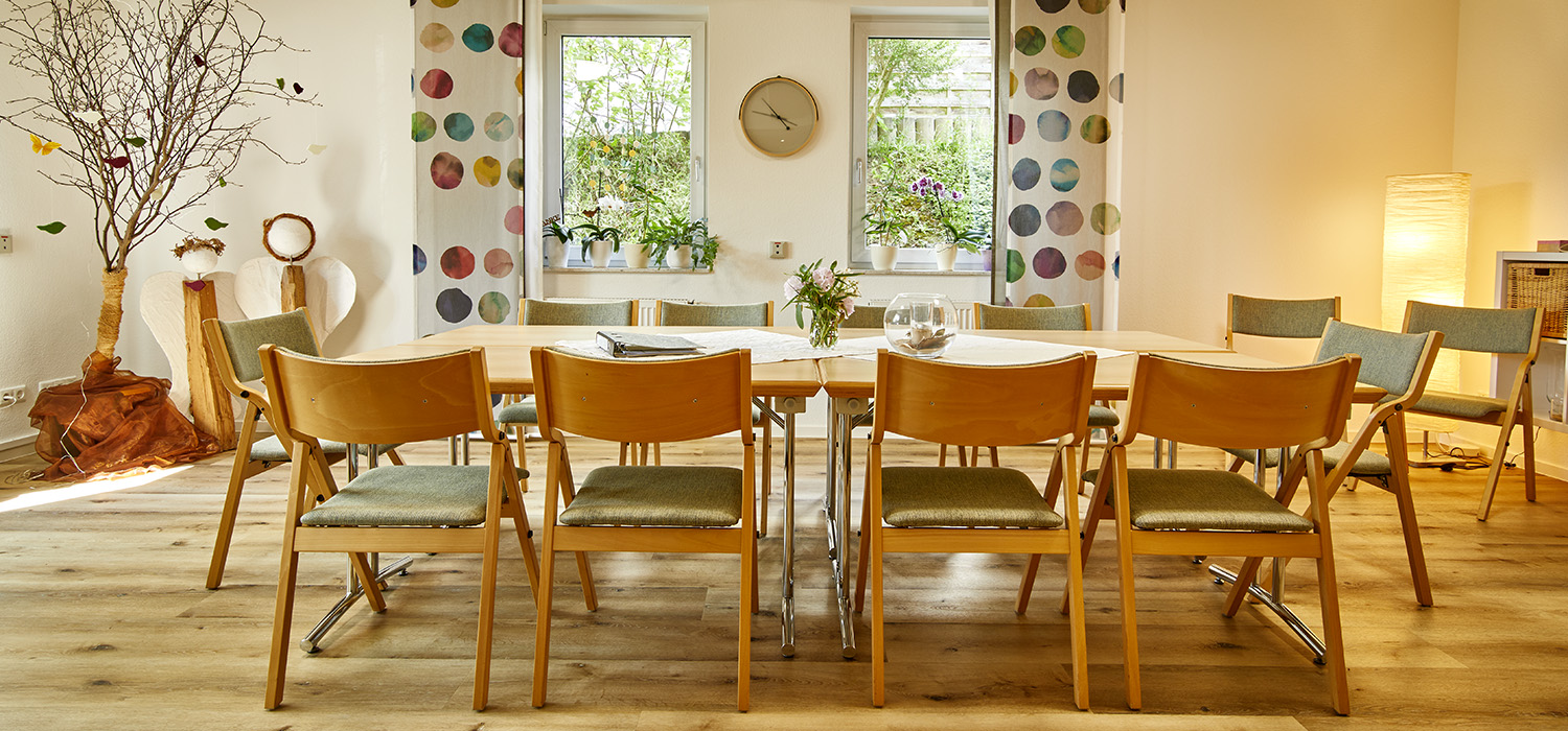 Gruppenraum mit Tisch, Holzstühlen, farbigen Vorhängen und Blick in den Garten