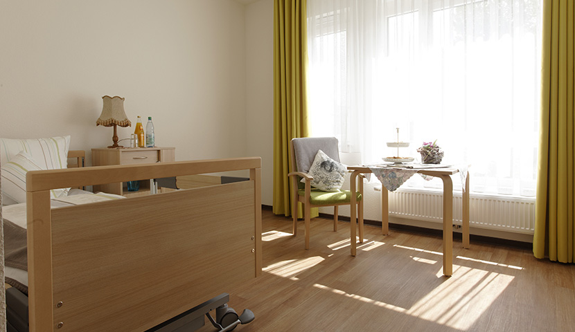 Bewohnerzimmer mit Bett, Stuhl, Tisch und bodentiefen Fenstern