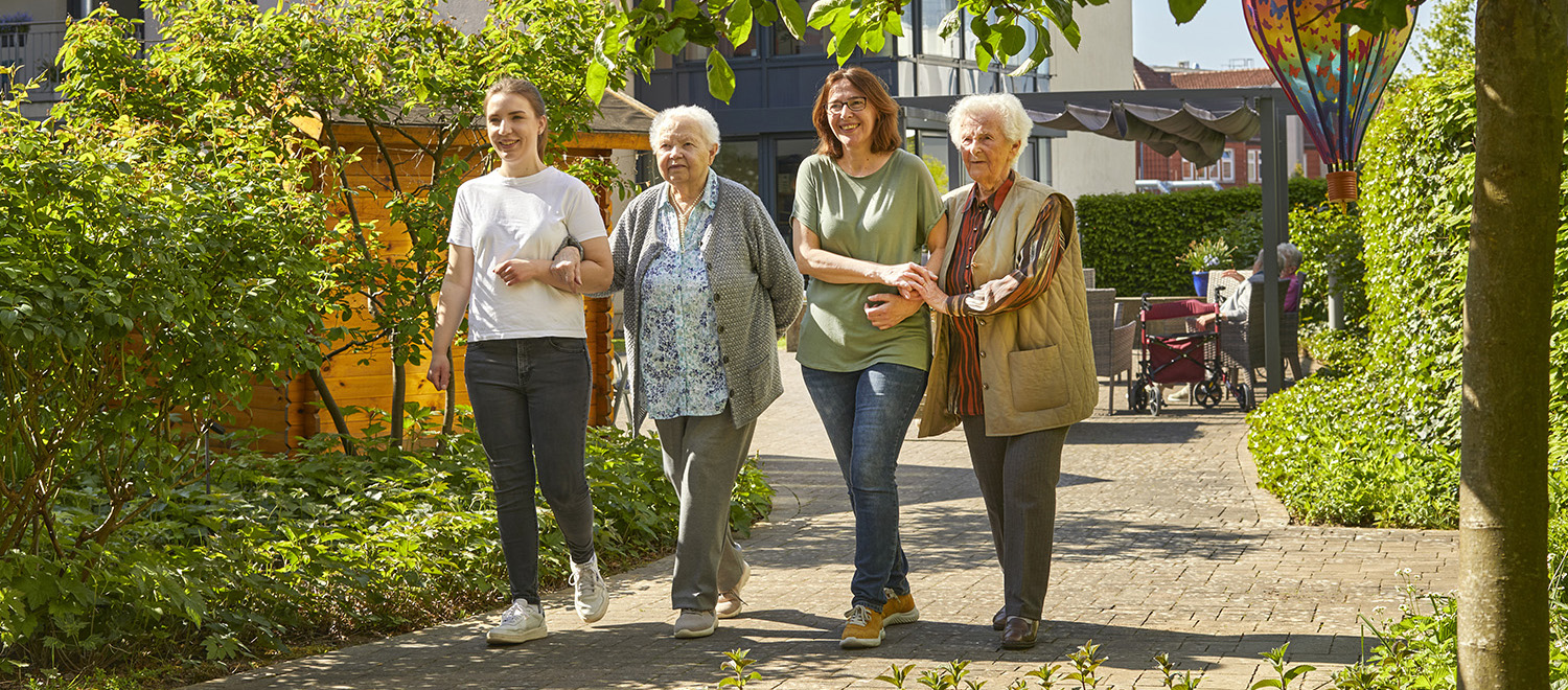 Zwei Tagespflegegästinnen gehen mit zwei Pflegekräften im Garten spazieren