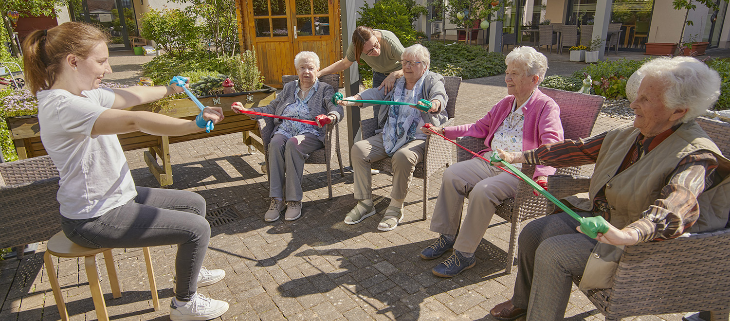 Vier Tagespflegegästinnen machen Bewegungsübungen im Garten unter Anleitung von zwei Pflegekräften