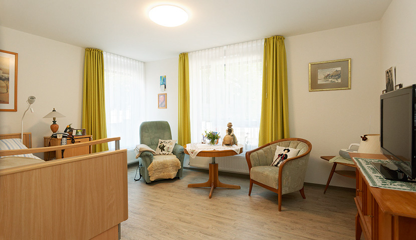 Helles Bewohnerzimmer mit Bett, zwei Sesseln, Kommode und großen Fenstern mit Gardinen